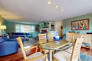 A living room with a glass-top dining table, wooden chairs, a blue sofa, a fireplace, and a bookshelf.