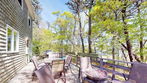 Una terraza de madera con mobiliario exterior, una parrilla y vistas a árboles.