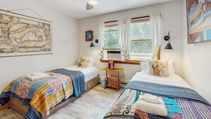 A bedroom with two beds, a ceiling fan, a window with curtains, a map on the wall, and a small table with a lamp.