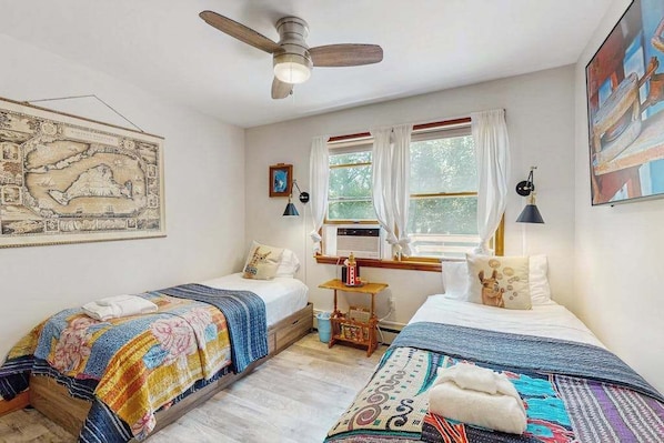 A bedroom with two beds, a ceiling fan, a window with curtains, a map on the wall, and a small table with a lamp.