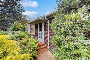 A house with a covered porch, surrounded by dense foliage and a variety of plants. - Welcoming island cottage with patio & enclosed yard - close to town & beaches (Edgartown)