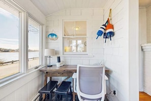 Een kamer met een houten bureau, een stoel, een raam met uitzicht op een strand, een lamp en nautische decoraties.