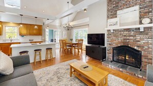 Uma sala de estar ampla com lareira, móveis de madeira e uma cozinha com armários de madeira e iluminação pendente.