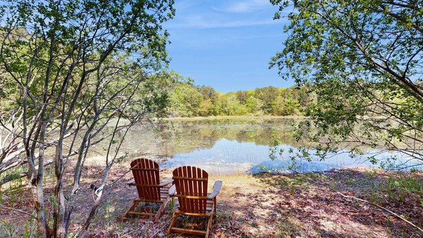 Duas cadeiras Adirondack à beira de um lago tranquilo, rodeado por árvores.