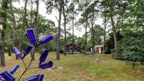 A backyard with a wooden cabin, a small shed, and a tree with blue bottles. - Comfy Cabin with Deck, Firepit, & Bike Path Access - Walk to Nauset Light Beach (Eastham)