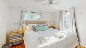 A bedroom with a bed, a ceiling fan, a nightstand, a lamp, and a window with curtains. - Peaceful waterview retreat with sunroom, AC, grill & amazing outdoor space (Eastham)