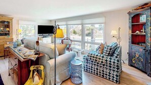 Een woonkamer met een bank, fauteuil en een televisie. Er is een boekenkast en een lamp.