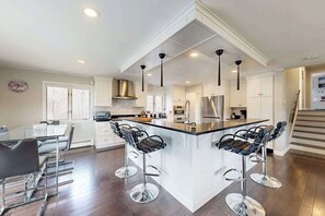 A modern kitchen with a central island, bar stools, a dining table, and a staircase. - Waterfront home with firepit, grill, open layout, smart TVs, deck, W/D, & AC (East Falmouth)