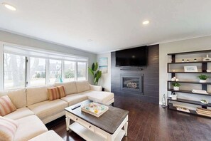 A living room with a sofa, coffee table, fireplace, TV, and shelves. - Waterfront home with firepit, grill, open layout, smart TVs, deck, W/D, & AC (East Falmouth)