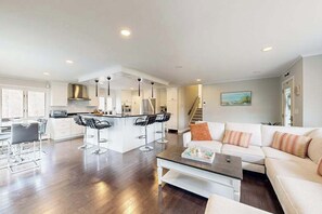 A modern living room with a sofa, coffee table, and a kitchen area with a bar. - Waterfront home with firepit, grill, open layout, smart TVs, deck, W/D, & AC (East Falmouth)