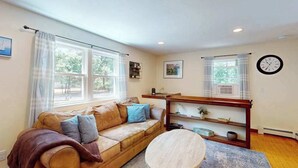 A living room with a sofa, a round coffee table, a clock, and a window with curtains. - Centrally located home near Nauset Light, Coast Guard Beach & Cape Rail Trail (Eastham)