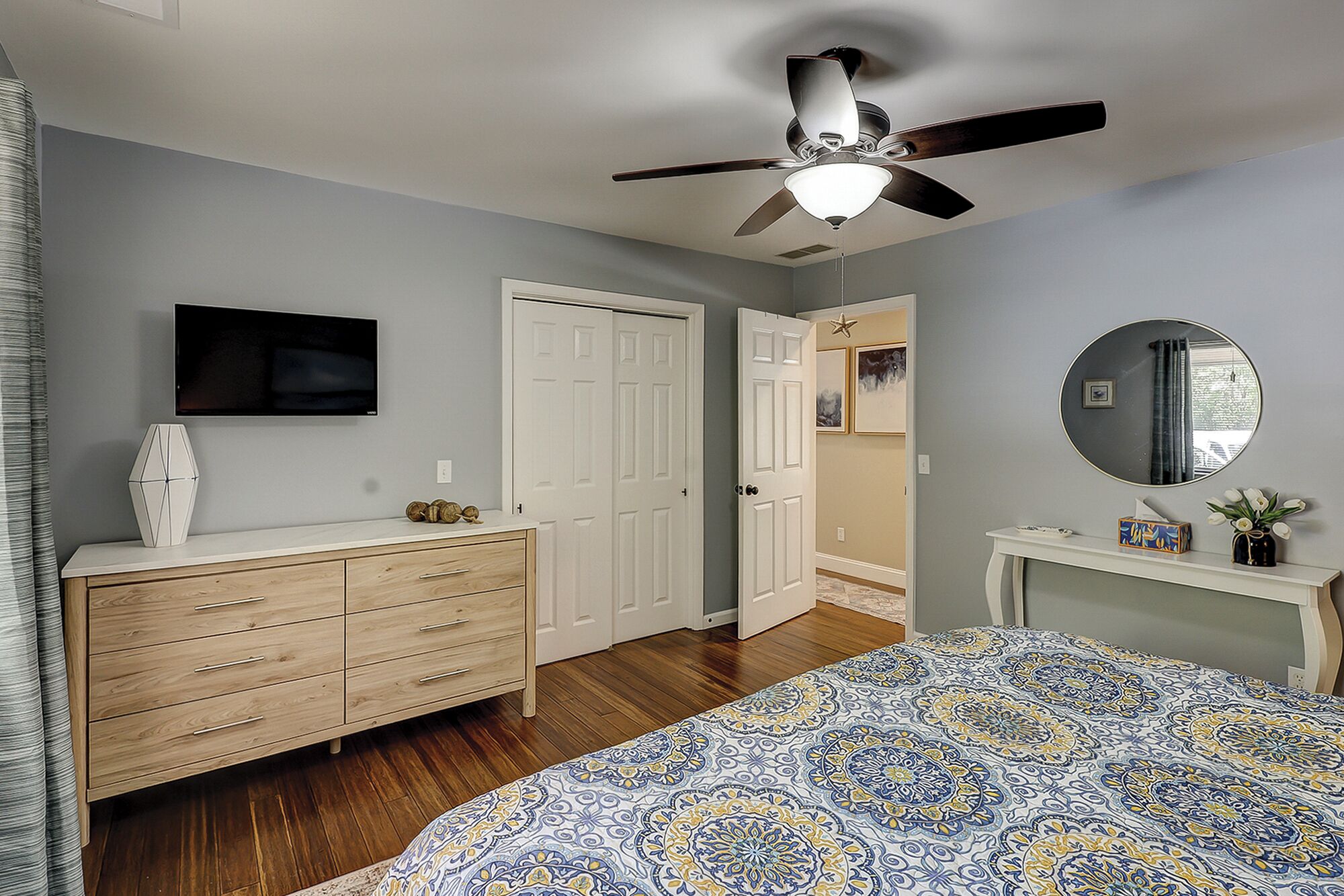 Une chambre à coucher comprenant un lit, une commode, un ventilateur de plafond, une télévision et une porte donnant sur une autre pièce.