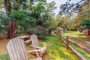 A wooden deck with Adirondack chairs and a picnic table, surrounded by trees and a wooden fence. - Charming, Dog-Friendly Cottage w/ Free WiFi & a Kitchenette - Close to the Beach (Eastham)