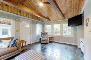 Ein Zimmer mit Holzbalkendecke, einem an der Wand montierten Fernseher, einer Couch mit gemusterten Kissen, einem Sessel und einem Fenster mit Blick ins Grüne.