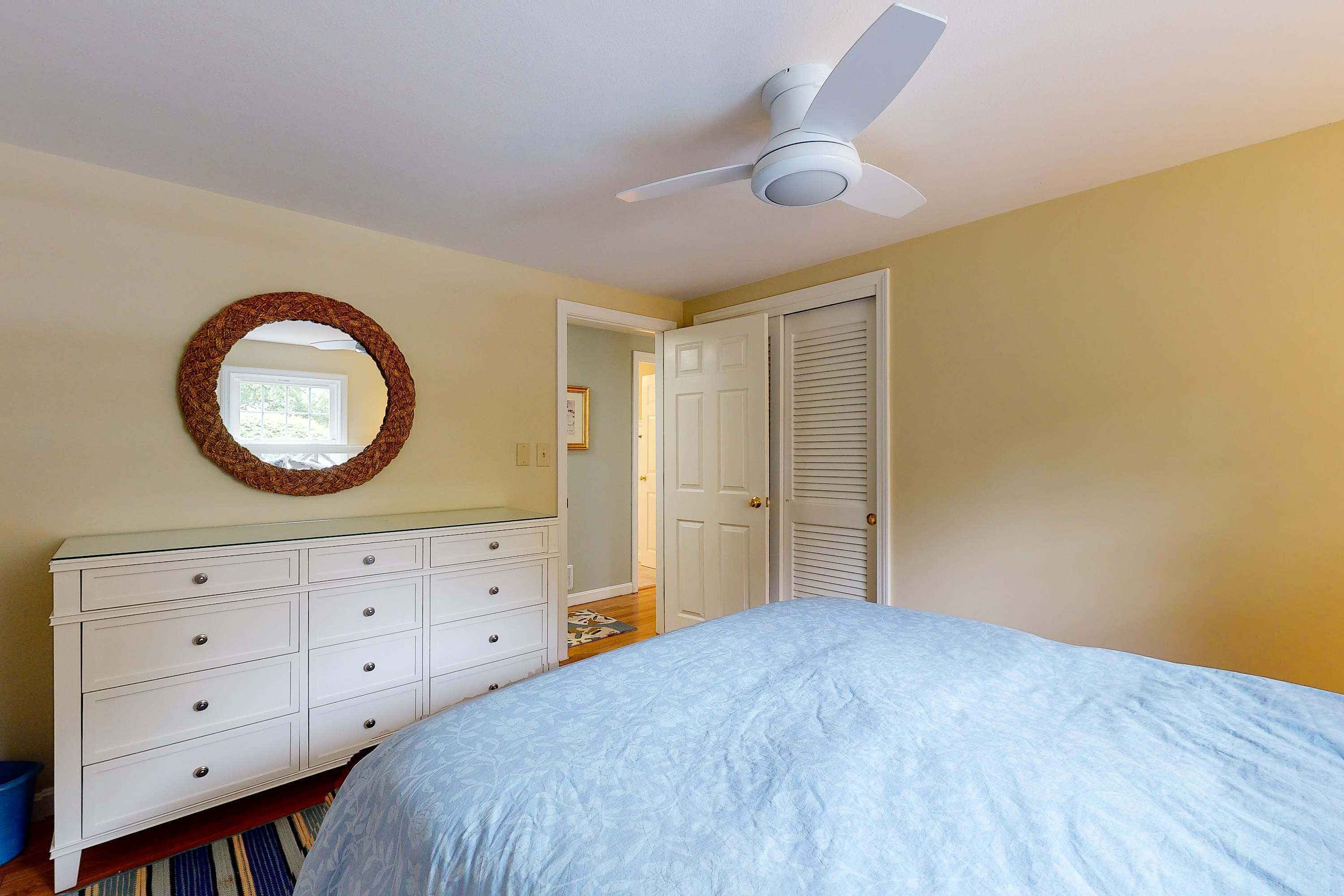 A bedroom with a bed, dresser, mirror, ceiling fan, and a door leading to another room.