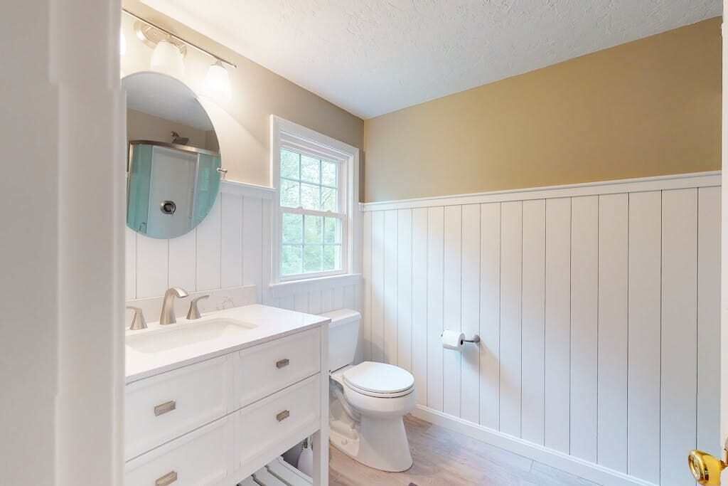 Un baño con tocador blanco, espejo redondo, inodoro y ventana.