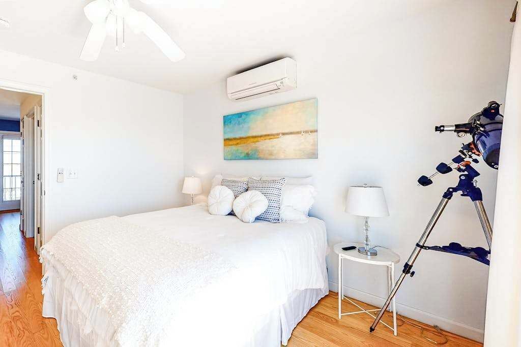 Un dormitorio con cama, mesita de noche, lámpara, aire acondicionado, un cuadro y un telescopio.
