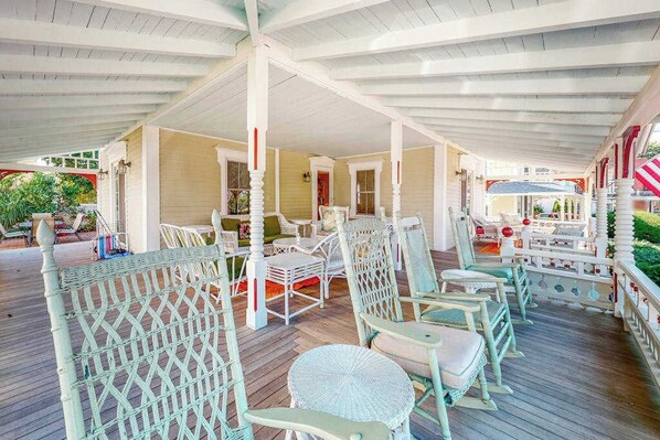 Een overdekt terras met rieten stoelen en tafels, een houten terras en een witte omheining.