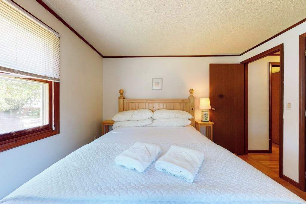 Una cama individual con cabecera, dos mesitas de noche con lámparas, una ventana con persianas y una puerta a otra habitación.