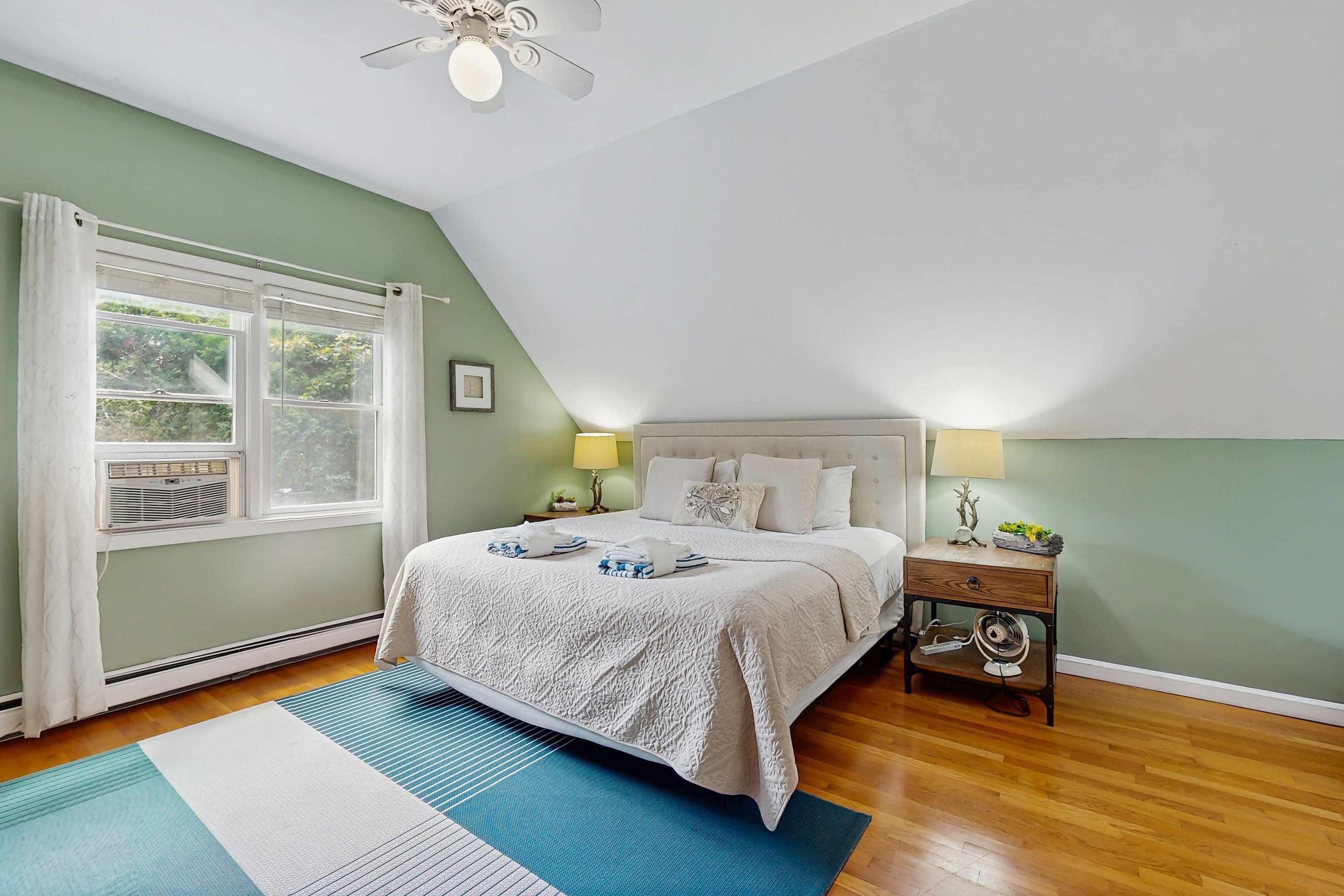 A bedroom with a bed, bedside lamps, a nightstand, a window with curtains, and a ceiling fan.