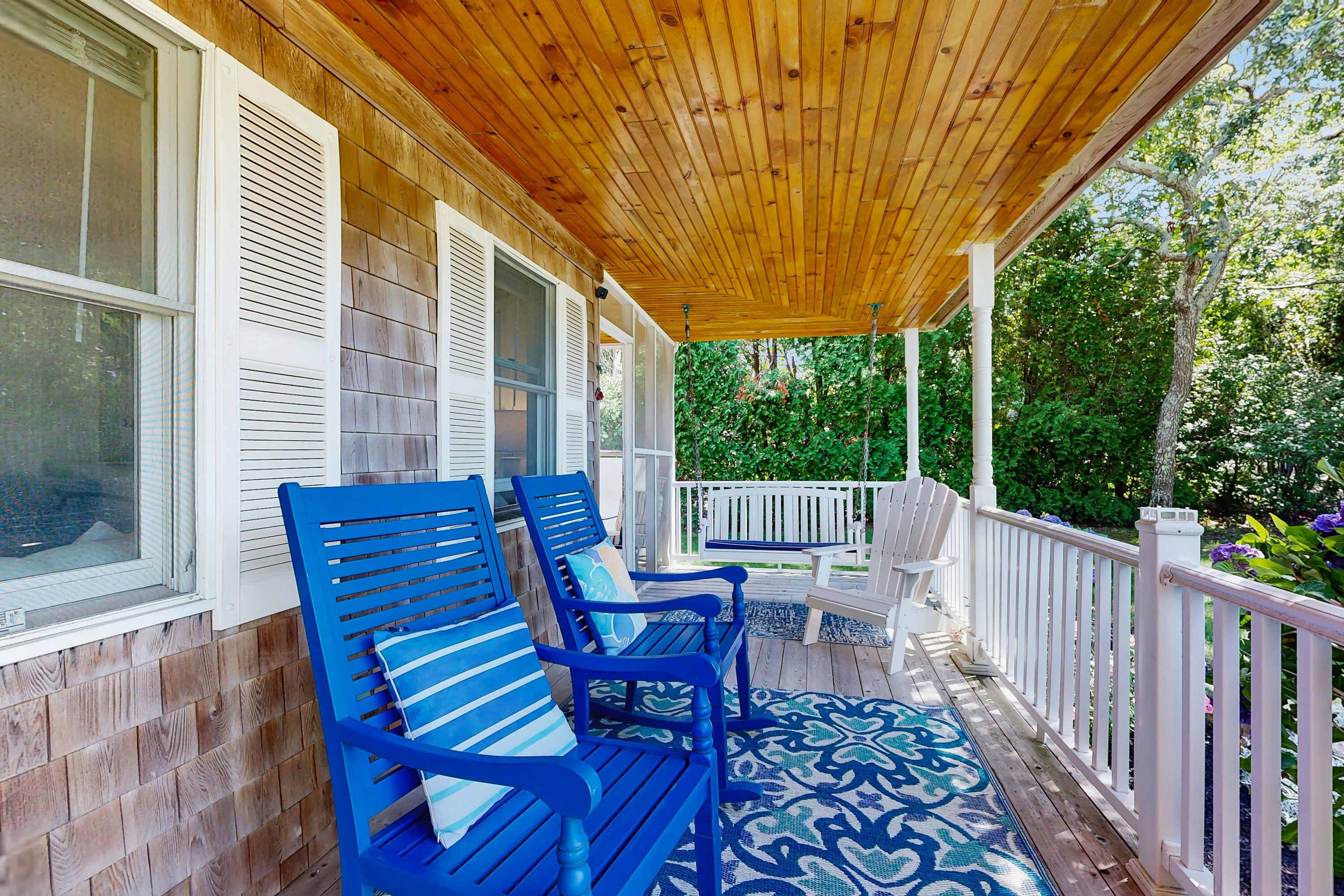 A covered porch with blue chairs and a white bench, wooden siding, and a view of greenery.