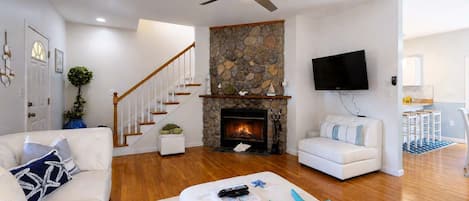A modern living room with a stone fireplace, white sofas, a ceiling fan, and a flat-screen TV.