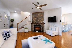 A modern living room with a stone fireplace, white sofas, a ceiling fan, and a flat-screen TV. - Newly remodeled home w/ new furniture, large yard & great porches (Oak Bluffs)