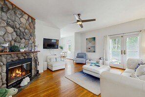 A modern living room with a stone fireplace, a flat-screen TV, and a ceiling fan. - Newly remodeled home w/ new furniture, large yard & great porches (Oak Bluffs)