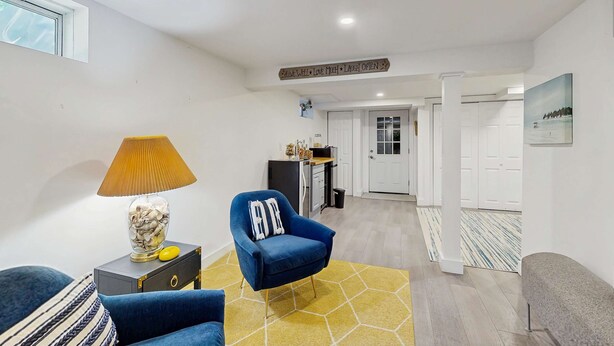 Un soggiorno moderno con una poltrona blu, una lampada e un contenitore di vetro decorativo. Un corridoio conduce a una cucina e a una porta.