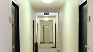 En smal, godt oplyst gang med døre på begge sider, en dørmåtte ved indgangen og et skilt over hovedet.