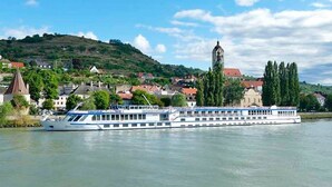 Ein Flusskreuzfahrtschiff mit mehreren Decks, ein markanter Turm im Hintergrund und eine Stadt mit Gebäuden und Grünflächen entlang des Flussufers.