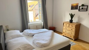 Ein ordentlich bezogenes Bett mit weißen Leinen, eine Holzkommode mit Schubladen, ein kleiner roter Hocker und ein Fenster mit Vorhängen.