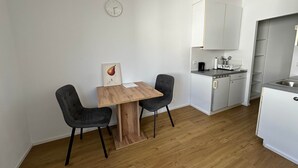 Une cuisine de taille réduite avec des meubles blancs, une table à manger en bois, deux chaises et une horloge au mur.