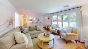 Ein Wohnzimmer mit einer Couch, Sesseln, einem Couchtisch und einem großen Fenster mit Vorhängen.