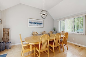 A dining area with a wooden table and chairs, a hanging light fixture, a framed artwork on the wall, and a window with blinds. - Golf course-front townhouse with private heated pool & poolside patio (Mashpee)