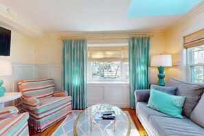 Ein Wohnzimmer mit gestreiften Sesseln, einem grauen Sofa, einem Glastisch und lindgrünen Vorhängen.