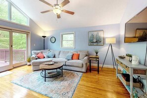 A living room with a sofa, coffee table, and a ceiling fan. There is a window, a clock, and a painting on the wall. - Dog-friendly home with great deck & association beach on Mashpee Pond (Mashpee)