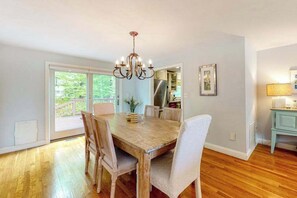 A dining area with a wooden table, chairs, a chandelier, and a view through glass doors to an outdoor deck. - Dog-friendly home with great deck & association beach on Mashpee Pond (Mashpee)
