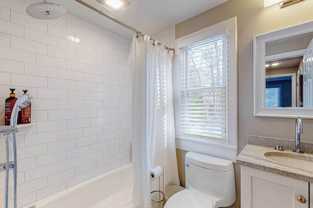 A bathroom with white tiles, a bathtub, a toilet, a showerhead, and a window with blinds.