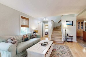 A living room with a sofa, coffee table, and a staircase leading to another room. - Serene wooded hideaway with spacious deck & Rail Trail access - near beach (Wellfleet)