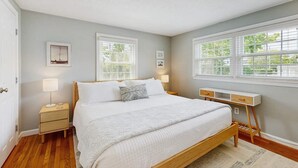 Quarto com cama, mesas de cabeceira, escrivaninha e duas janelas.