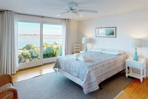 Une chambre à coucher avec un lit, un ventilateur de plafond, une fenêtre donnant sur un beau paysage, une chaise et une table de chevet avec une lampe.