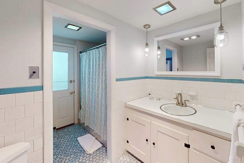 Une salle de bain avec un lavabo blanc, un miroir et des armoires, une douche avec un sol carrelé en bleu et blanc, et une porte donnant sur une autre pièce.