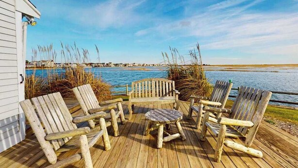 Eine Holzterrasse mit Adirondack-Stühlen und einem kleinen Tisch, mit Blick auf ein Gewässer und eine entfernte Küste.