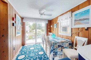 A room with wooden walls, a ceiling fan, a table with a blue tablecloth, and a view of the outdoors. - Secluded ranch home with a full outdoor shower & private community beach (Brewster)