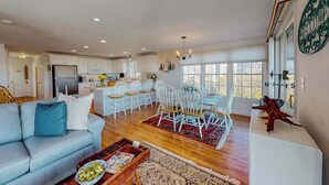 A spacious living area with a sofa, dining table, and chairs, a kitchen in the background, and large windows. - Private water access & stunning views (Barnstable)