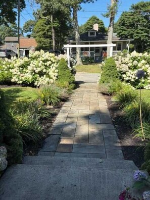 Un chemin pavé mène à une maison dotée d’un porche, qui est entourée de plantes à fleurs blanches et de verdure.