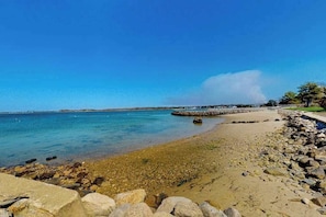 Uma praia com águas cristalinas, costa rochosa e um cais ao longe.