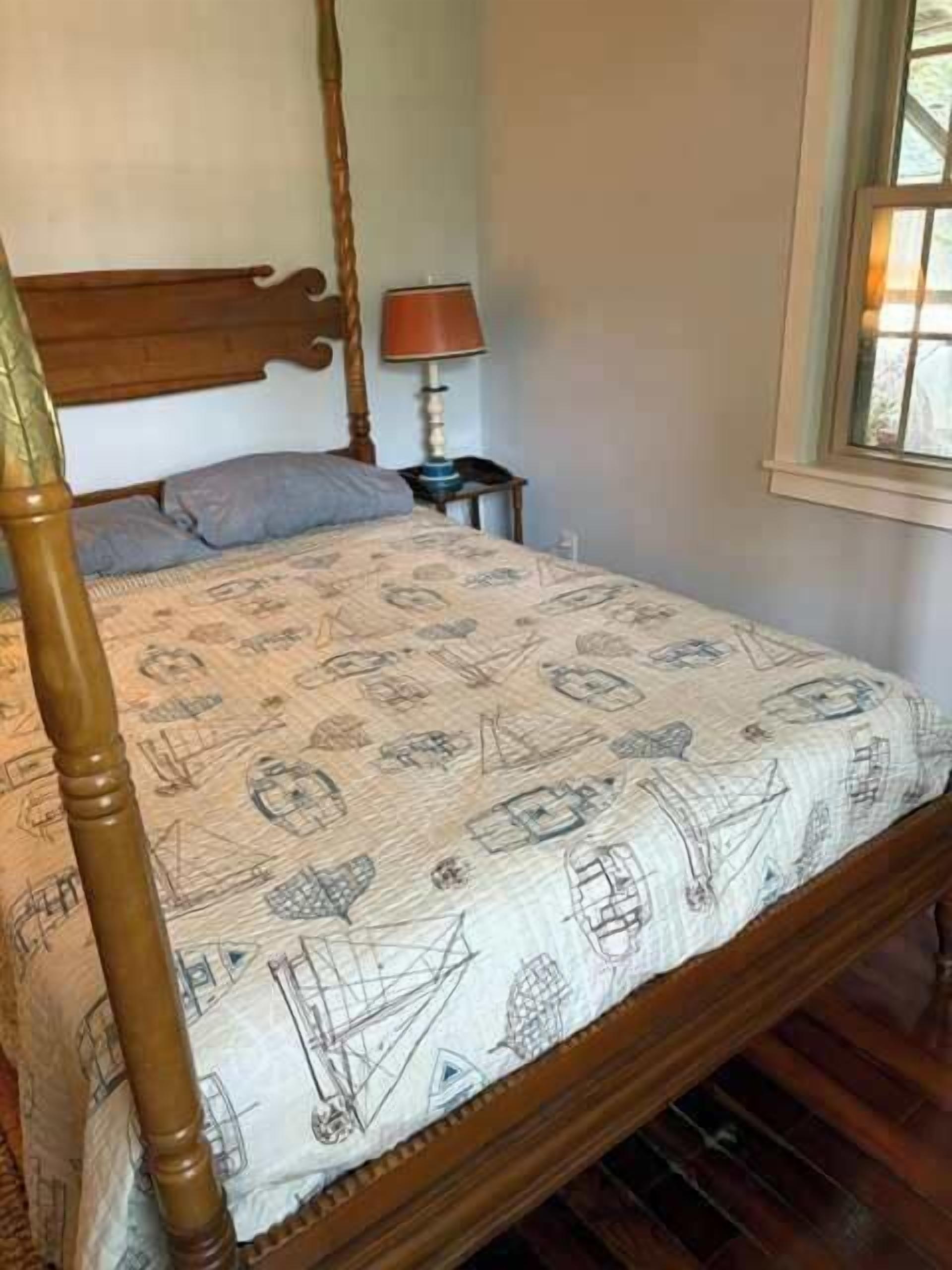A bed with a patterned bedspread, a wooden headboard, a nightstand with a lamp, and a window with daylight.