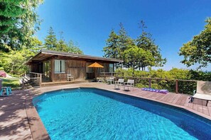A swimming pool with a wooden deck, a small wooden building with a roof, and outdoor seating.